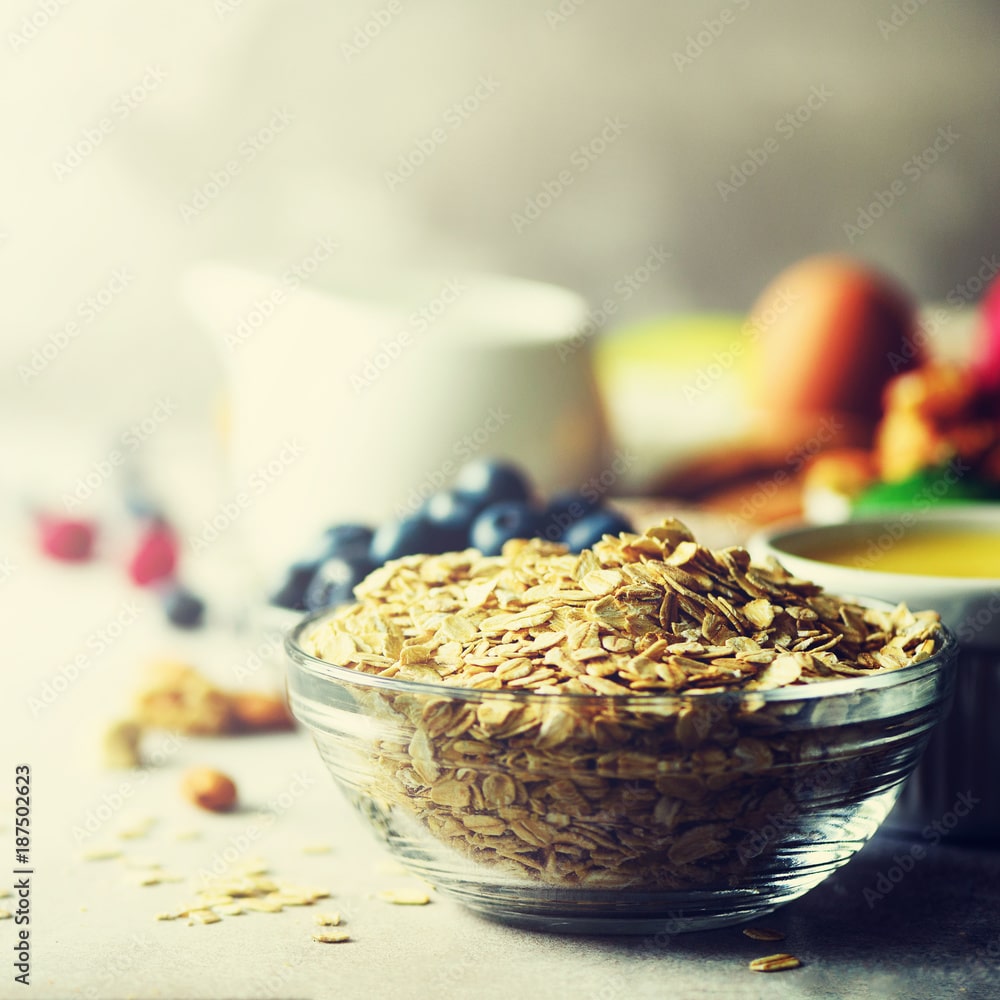 Bol de flocons d'avoine devant un pichet de lait, des myrtilles et des fruits rouges pour un petit-déjeuner sain. Adobe Stock | 187502623.