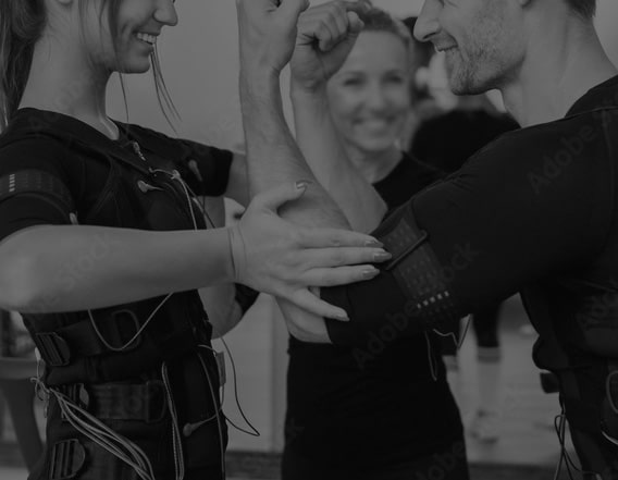 Couple souriant en tenue EMS (stimulation électrique musculaire) ajustant leurs bras pour un exercice, une entraîneuse sourit en arrière-plan.