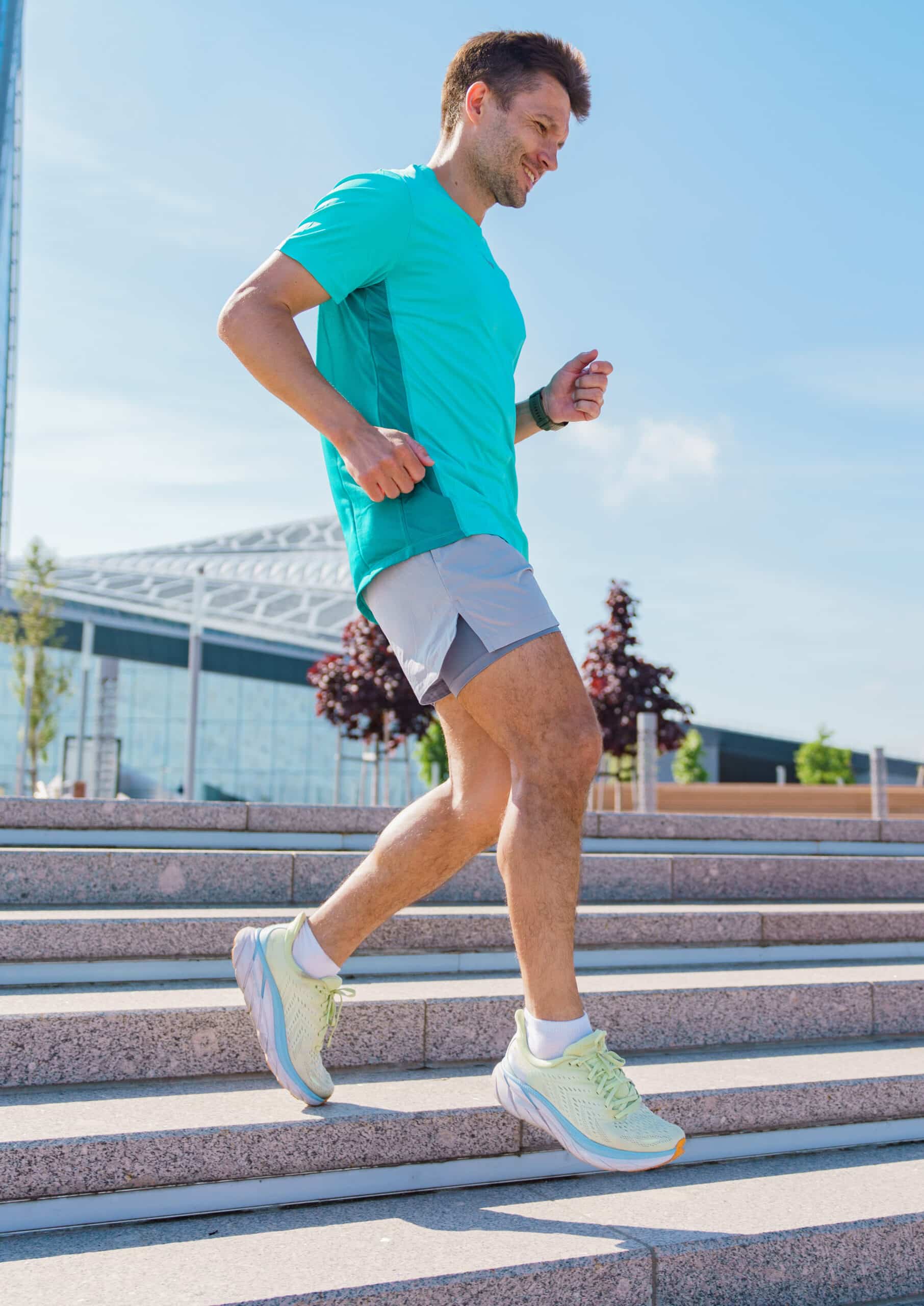 Homme souriant en tenue de sport turquoise et short gris montant des escaliers en granit par un jour ensoleillé.