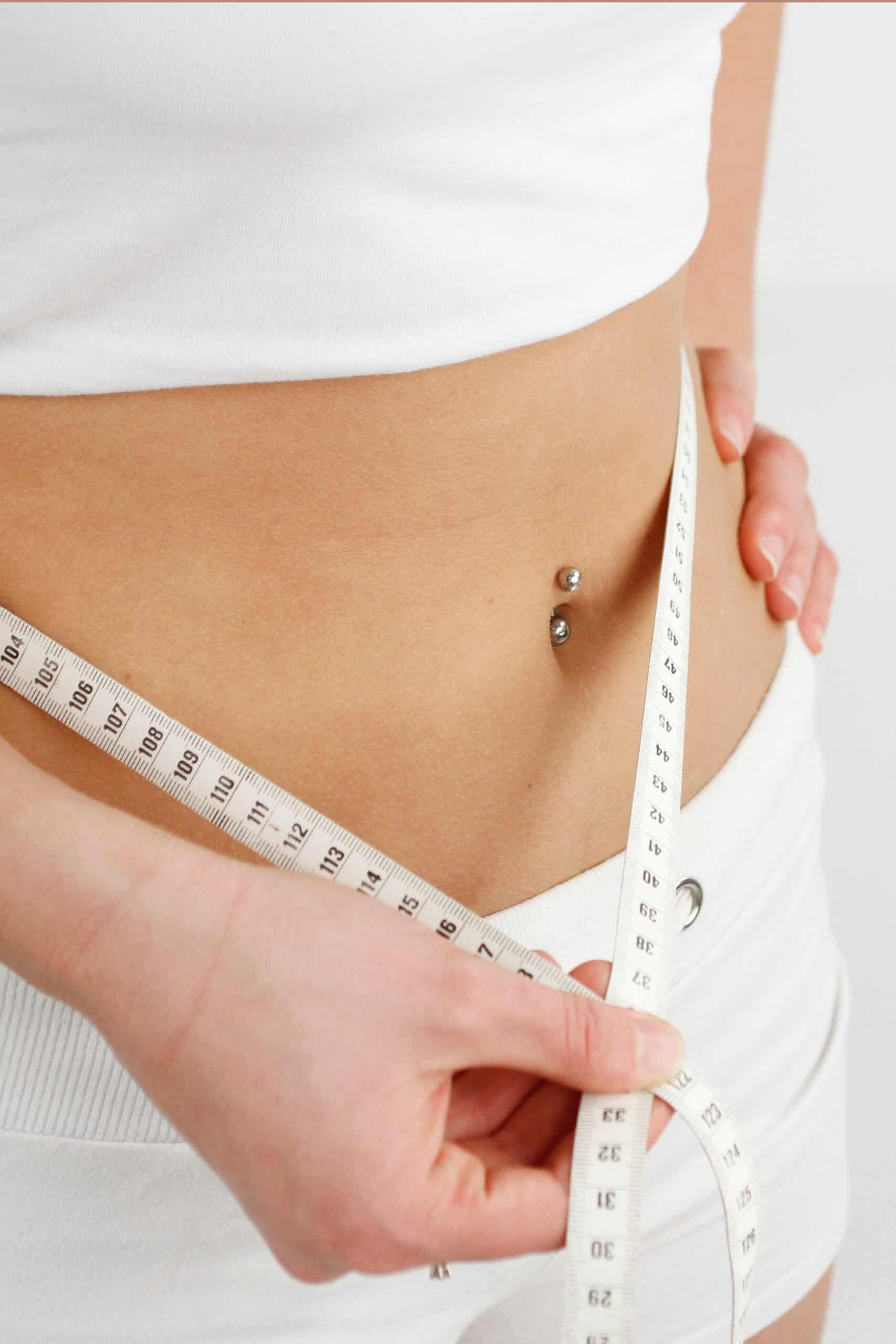 Femme mesurant sa taille avec un mètre ruban blanc autour du ventre, affichant des chiffres comme 104 à 114 et 29 à 33.